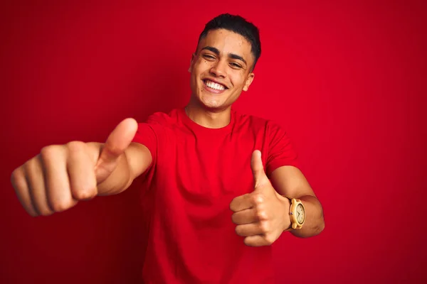 孤立した赤い背景の上に立つTシャツを着た若いブラジル人男性は 手でポジティブなジェスチャーを行い 親指を立てて笑顔を浮かべて成功を収めました 勝者のジェスチャー — ストック写真