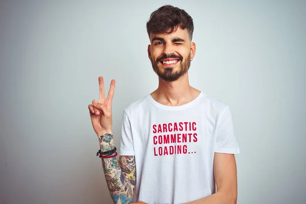 Young man with tattoo wearing fanny t-shirt standing over isolated white background smiling with happy face winking at the camera doing victory sign. Number two.