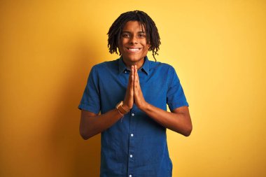 Rahat kot gömlek giyen dreadlocks ile Afro adam elleri birlikte emin gülümseyerek af isteyen izole sarı arka plan üzerinde ayakta.