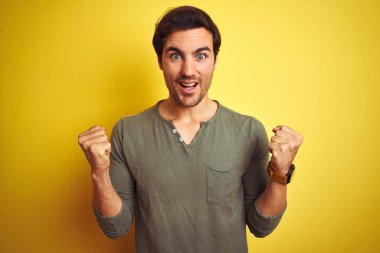 İzole edilmiş sarı arka planda dikilmiş tişört giyen genç yakışıklı adam kollarını kaldırarak ve açık gözlerle başarıyı kutluyor ve şaşırıyor. Kazanan konsept.