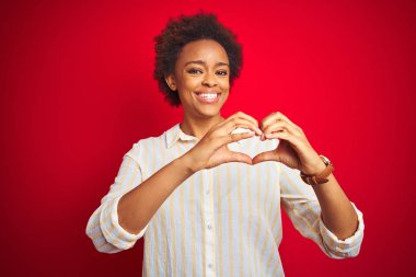 Izole kırmızı arka plan üzerinde afro saçlı genç güzel afro-amerikan kadın aşk kalp sembolü ve elleriyle şekil gösteren gülümseyerek. Romantik kavram.