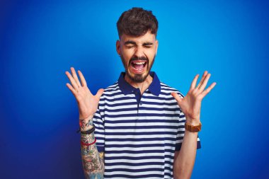 Çizgili polo giyen dövmegiyen genç adam izole mavi arka plan üzerinde duran deli ve heyecan çığlık kapalı gözleri ile başarı için deli ve deli kutluyor. Kazanan kavramı
