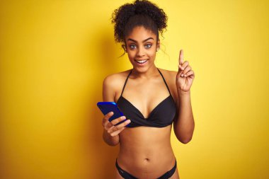 Izole sarı arka plan üzerinde akıllı telefon kullanarak bikini giyen Afro-Amerikan kadın mutlu yüz, bir numaralı ile bir fikir veya soru işaret parmağı ile şaşırttı