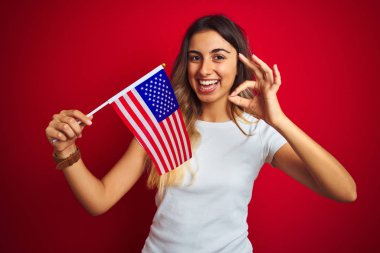 Kırmızı izole arka plan üzerinde Amerika bayrağı tutan genç kadın parmakları ile ok işareti yapıyor, mükemmel sembolü