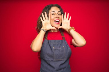 Kırmızı izole arka plan üzerinde önlük üniforma giyen orta yaş yaşlı kadın Gülümseyen neşeli oynarken yüzleri gösteren elleri ile bir boo gözetleme. Şaşırttı ve çıktı