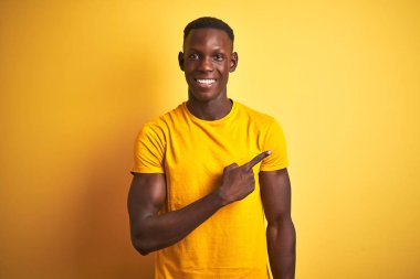 Genç afro-amerikan adam rahat ve doğal ifade ile yan yana el ve parmak ile işaret yüzünde bir gülümseme ile neşeli izole sarı arka plan üzerinde duran rahat t-shirt giyen