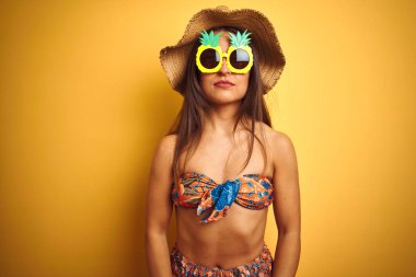 Bikini ve ananas güneş gözlüğü takan tatilde olan kadın, yüzünde ciddi bir ifade ile izole sarı arka plan üzerinde. Kameraya basit ve doğal bir bakış.