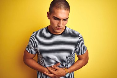 Genç yakışıklı adam mide üzerinde el ile sarı izole arka plan üzerinde çizgili t-shirt giyiyor çünkü hazımsızlık, ağrılı hastalık kötü hissediyorum. Ağrı kavramı.