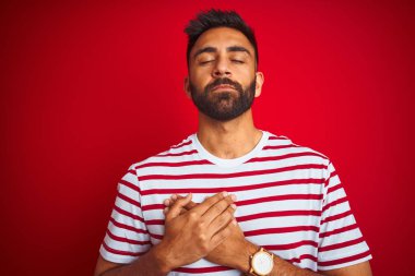 İzole kırmızı arka plan üzerinde duran çizgili t-shirt giyen genç hintli adam kapalı gözleri ve yüzünde minnettar jest ile göğüs üzerinde gülümseyerek. Sağlık kavramı.