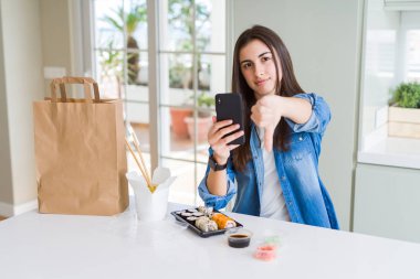 Beautiful young woman ordering food delivery from app using smartphone with angry face, negative sign showing dislike with thumbs down, rejection concept clipart