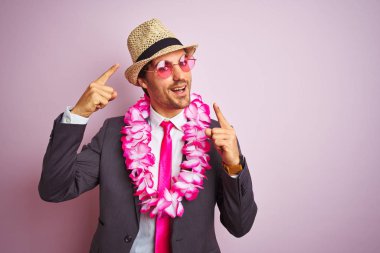 Takım elbise şapka gözlük hawaiian lei giyen genç işadamı izole pembe arka plan üzerinde çok mutlu el ve parmak ile işaret