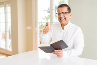 Orta yaş adam evde bir kitap okuma gözlük büyük gülümseme ok işareti yapıyor mutlu giyen, parmakları ile başparmak, mükemmel işareti