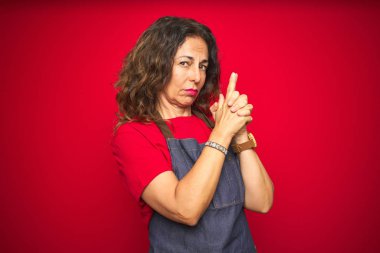 Orta yaş yaşlı kadın kırmızı izole arka plan üzerinde önlük üniforma giyen el hareketi ile sembolik silah holding, çekim silah öldürme oynarken, öfkeli yüz