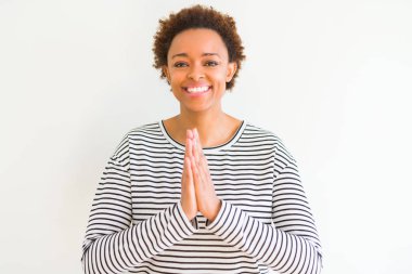 Genç güzel afro-amerikan kadın beyaz arka plan üzerinde çizgili kazak giyen elleri birlikte emin gülümseyerek af isteyen dua.