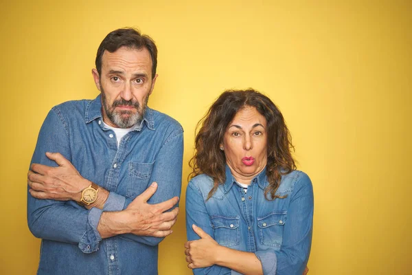 Güzel orta yaş çift birlikte izole sarı arka plan sallayarak üzerinde duran ve yüzünde üzgün ve şok ifade ile kış soğuk için donma