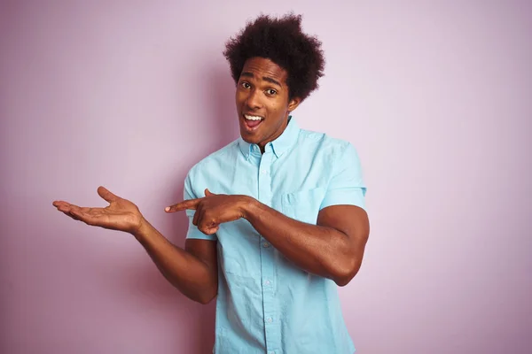 Young american man with afro hair wearing blue shirt standing over isolated pink background amazed and smiling to the camera while presenting with hand and pointing with finger.