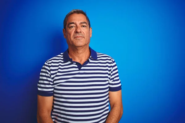 Handsome middle age man wearing striped polo standing over isolated blue background with serious expression on face. Simple and natural looking at the camera.