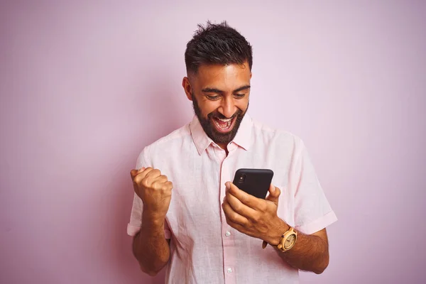 Young indian man using smartphone standing over isolated pink background screaming proud and celebrating victory and success very excited, cheering emotion