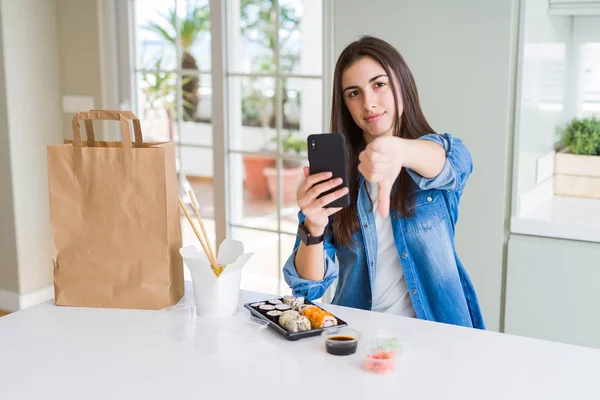 Beautiful young woman ordering food delivery from app using smartphone with angry face, negative sign showing dislike with thumbs down, rejection concept