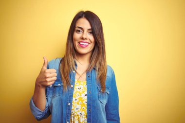 Genç güzel kadın el ile jest kadar mutlu başparmak yapıyor sarı izole arka plan üzerinde duran. Başarı gösteren kameraya bakarak ifadeyi onaylama.