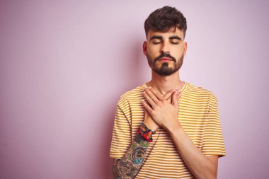 Sarı çizgili t-shirt giyen dövme giyen genç adam kapalı gözleri ve yüzünde minnettar jest göğüs elleri ile gülümseyerek izole pembe arka plan üzerinde duran. Sağlık kavramı.