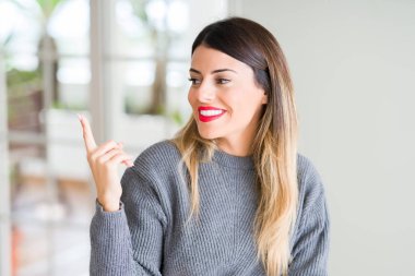 Kameraya bakarak yan el ve parmak ile işaret yüzünde, evde kocaman bir gülümseme ile kış kazak giyen genç güzel kadın.