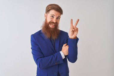 Genç kızıl saçlı İrlandalı işadamı izole beyaz arka plan üzerinde duran takım elbise giyen mutlu yüz zafer işareti yapıyor kameraya göz kırparak gülümseyerek. İki numara.