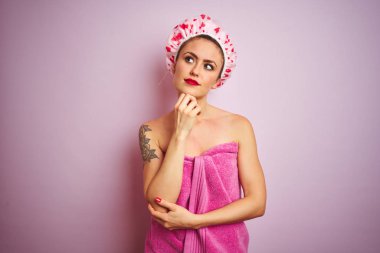 Havlu ve banyo şapkası takan genç güzel kadın duştan sonra pembe izole arka plan üzerinde el ile çeneüzerinde düşünme soru, dalgın ifade. Düşünceli bir yüzle gülümsüyor. Şüphe kavramı.
