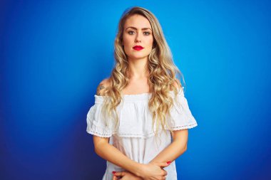 Yüzünde ciddi bir ifade ile mavi izole arka plan üzerinde duran beyaz elbise giyen genç güzel kadın. Kameraya basit ve doğal bir bakış.