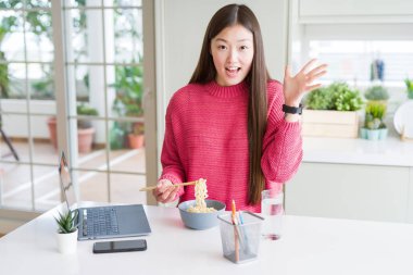 Güzel Asyalı kadın dizüstü bilgisayar kullanarak çalışan ve asya erişte yeme çok mutlu ve heyecanlı, kazanan ifade büyük bir gülümseme ile çığlık zafer kutluyor ve ellerini kaldırdı