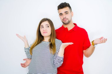 Genç güzel çift birlikte beyaz izole arka plan üzerinde habersiz ve kollar ve eller kaldırdı ile karışık ifade. Şüphe kavramı.