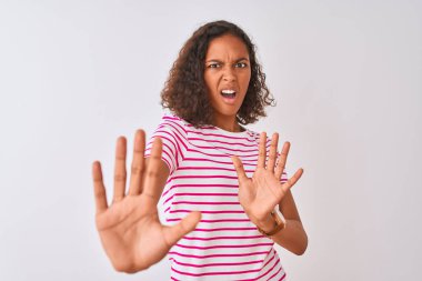 İzole beyaz arka plan üzerinde duran pembe çizgili t-shirt giyen brezilyalı genç kadın korku ve korku ile dehşete elleriyle jest durdurmak, şok içinde bağırarak. Panik kavramı.