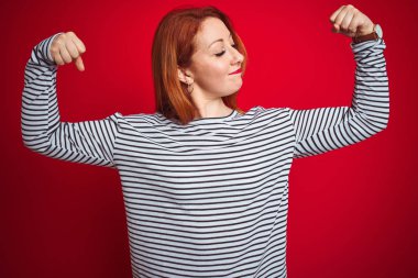 Genç kızıl saçlı kadın kayışlar lacivert gömlek giyen kırmızı izole arka plan üzerinde duran kol kasları gururla gülümseyen gösteren. Fitness konsepti.