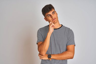 Lacivert çizgili tişört giyen genç hintli adam izole beyaz arka plan üzerinde duran elleri çeneüzerinde soru, dalgın ifade düşünüyor. Düşünceli bir yüzle gülümsüyor. Şüphe kavramı.