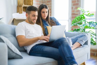 Genç çift yeni bir ev bilgisayar laptop kullanarak kanepede oturan rahatlatıcı, yeni daireye taşımak için mutlu gülümseyen hareket