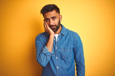İzole sarı arka plan üzerinde duran denim gömlek giyen genç hintli adam yorgun görünümlü ve çapraz kolları ile depresyon sorunları ile sıkılmış düşünme.