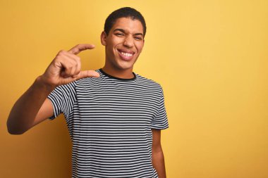 Genç yakışıklı Arap adam izole sarı arka plan üzerinde lacivert çizgili t-shirt giyen gülümseyerek ve emin el parmakları görünümlü ve kamera ile küçük boyutlu işareti yapıyor gesturing. Ölçü kavramı.