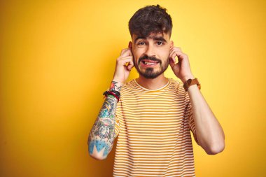 Dövme li genç adam yüksek sesle müzik sesi için rahatsız ifade ile parmakları ile kulakları kapsayan izole sarı arka plan üzerinde duran çizgili t-shirt giyiyor. Sağır kavramı.