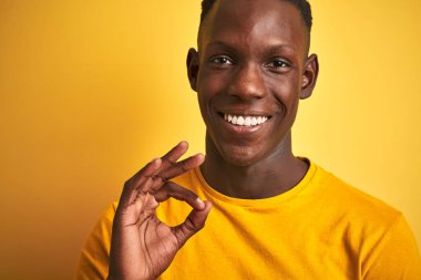 Izole sarı arka plan üzerinde duran rahat t-shirt giyen Afrikalı Amerikalı adam parmakları ile ok işareti yapıyor, mükemmel sembolü