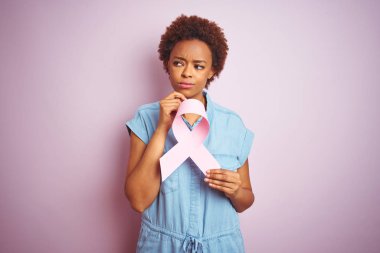 Genç afro-amerikan kadın izole pembe arka plan üzerinde brest kanser kurdele tutan ciddi yüz soru hakkında düşünme, çok karışık bir fikir