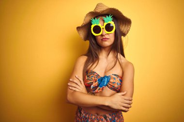 Bikini ve ananas güneş gözlüğü takan tatilde olan kadın izole sarı arka plan üzerinde şüpheci ve sinirli, çapraz kolları ile yüzünde onaylamayan ifade. Negatif kişi.