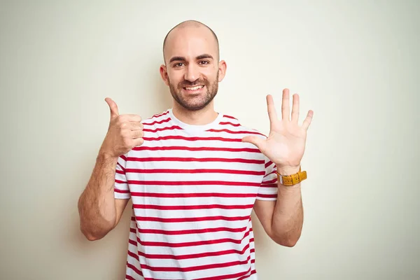 白い孤立した背景の上にカジュアルなストライプの赤いTシャツを着たあごひげの若いハゲ男は 自信を持って幸せに微笑みながら 指番号6を示し 指を上げる — ストック写真