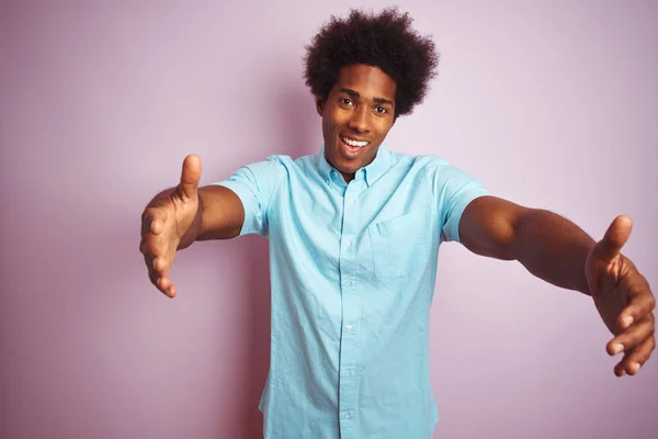 年轻的美国男子 一个非洲的头发 穿着蓝色衬衫站在孤立的粉红色背景 看着相机微笑着张开双臂拥抱 拥抱幸福的欢快表达 — 图库照片