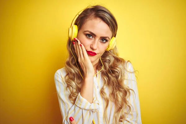 黄色の孤立した背景の上にヘッドフォンを使用して音楽を聴く若い美しい女性は 腕を組んだうつ病の問題に疲れて退屈しているように見えます — ストック写真