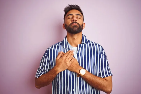 Young handsome indian man wearing summer striped shirt over isolated pink background smiling with hands on chest with closed eyes and grateful gesture on face. Health concept.