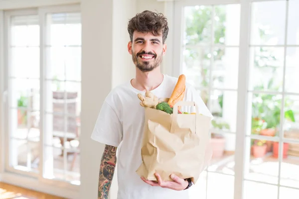 Young man holding paper bag of fresh groceries from the supermarket with a happy face standing and smiling with a confident smile showing teeth