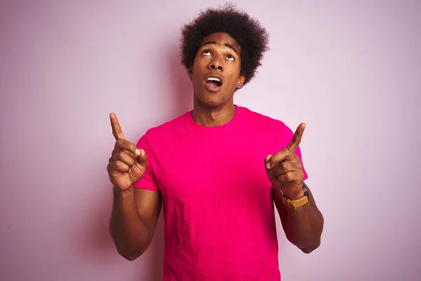 孤立したピンクの背景の上に立つTシャツを着たアフロヘアの若いアメリカ人男性は驚き 指で指を指差し 腕を上げて驚いた — ストック写真