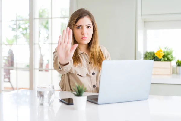 Beautiful young woman using computer laptop with open hand doing stop sign with serious and confident expression, defense gesture