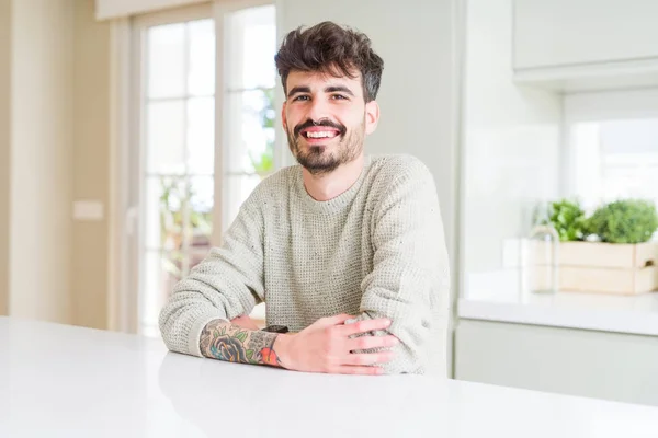 Young man wearing casual sweater sitting on white table happy face smiling with crossed arms looking at the camera. Positive person.