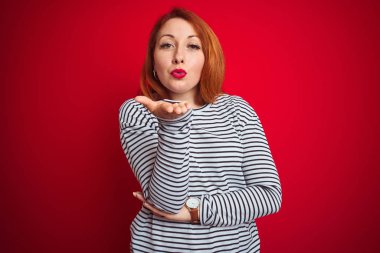 Genç kızıl saçlı kadın kayışlar lacivert gömlek kırmızı izole arka plan üzerinde duran kamera hava güzel ve seksi olmak el iyle bir öpücük üfleme bakarak giyiyor. Aşk ifadesi.